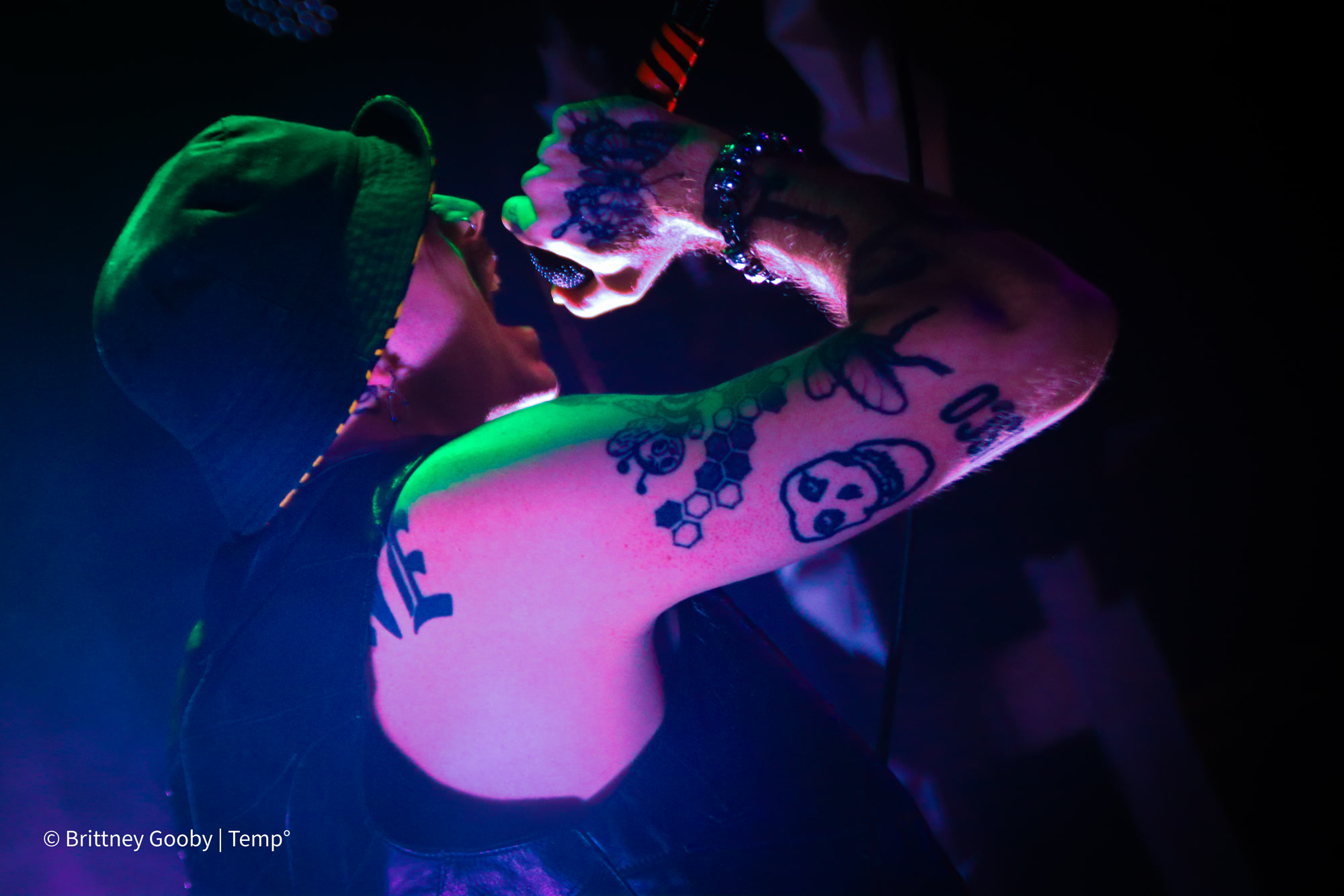 Blood Orchid sings into a microphone in a green bucket hat, arms raised under green and magenta light