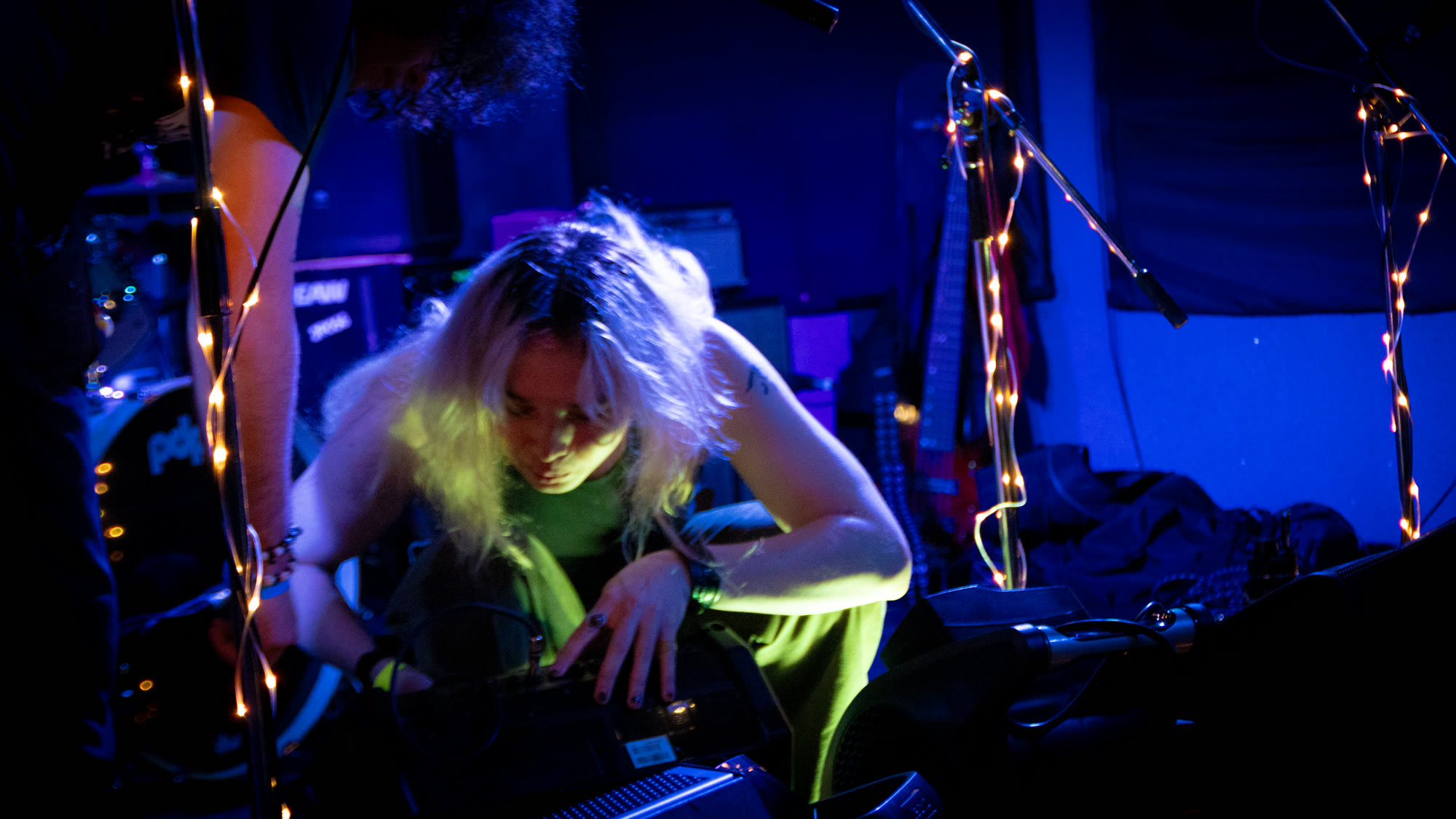 Musician with light hair leans over a pedalboard under blue light and string lights