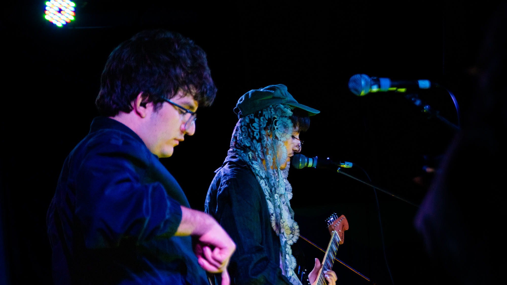 Two musicians perform, one in glasses and one in a lace head covering playing guitar
