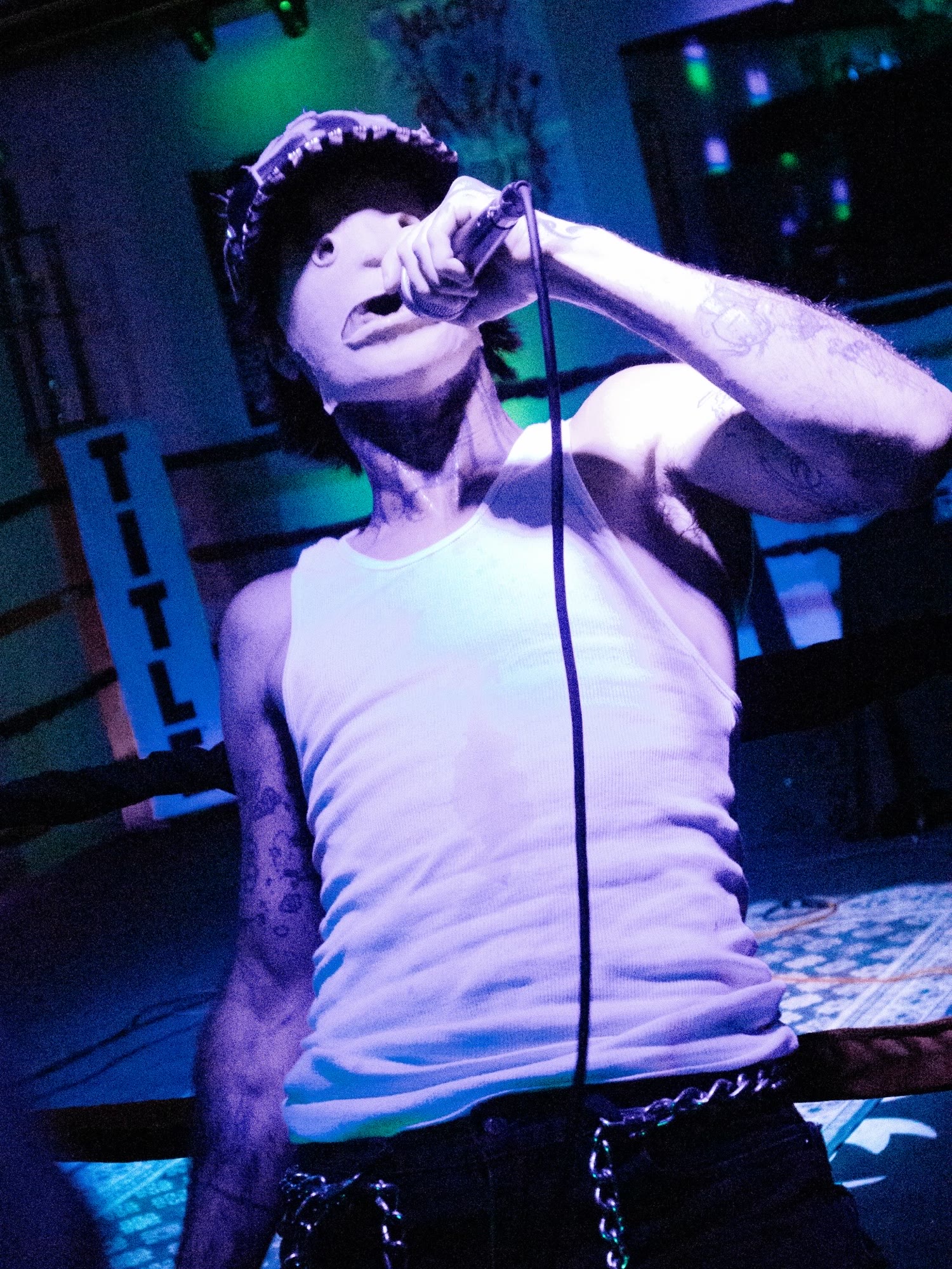 Singer in a white tank top and studded cap sings into a microphone under purple light