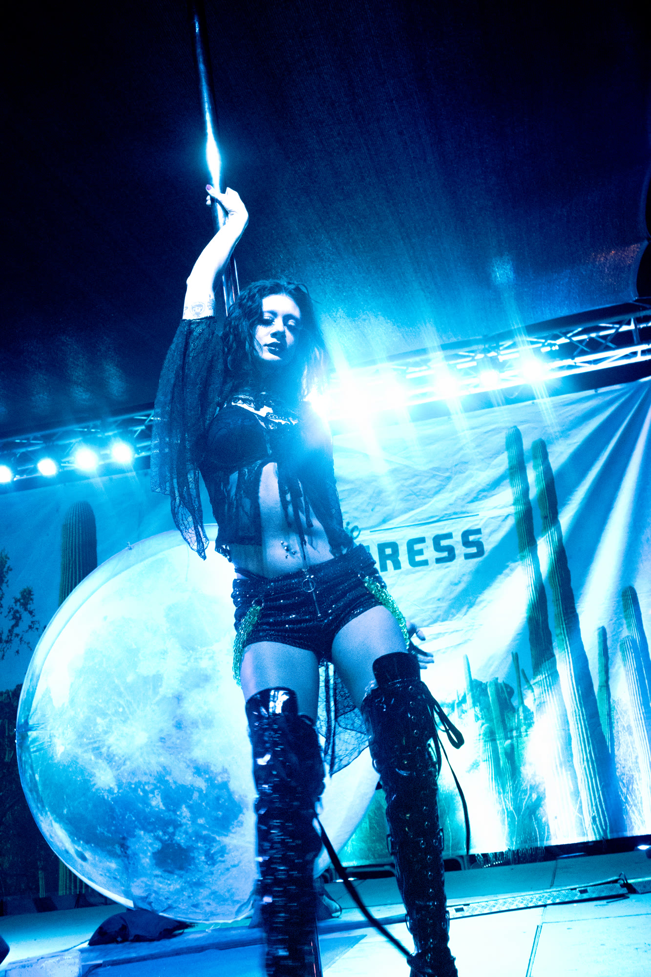 Performer holds a pole on an outdoor stage under blue light, with a moon and cactus backdrop