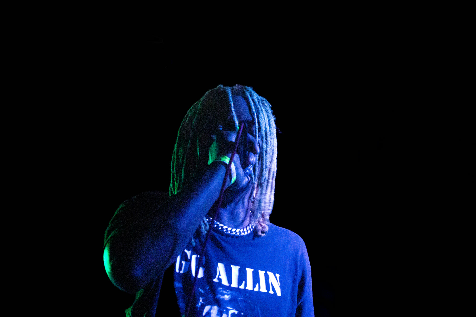 Performer with long dreadlocks sings into a microphone under blue and green light