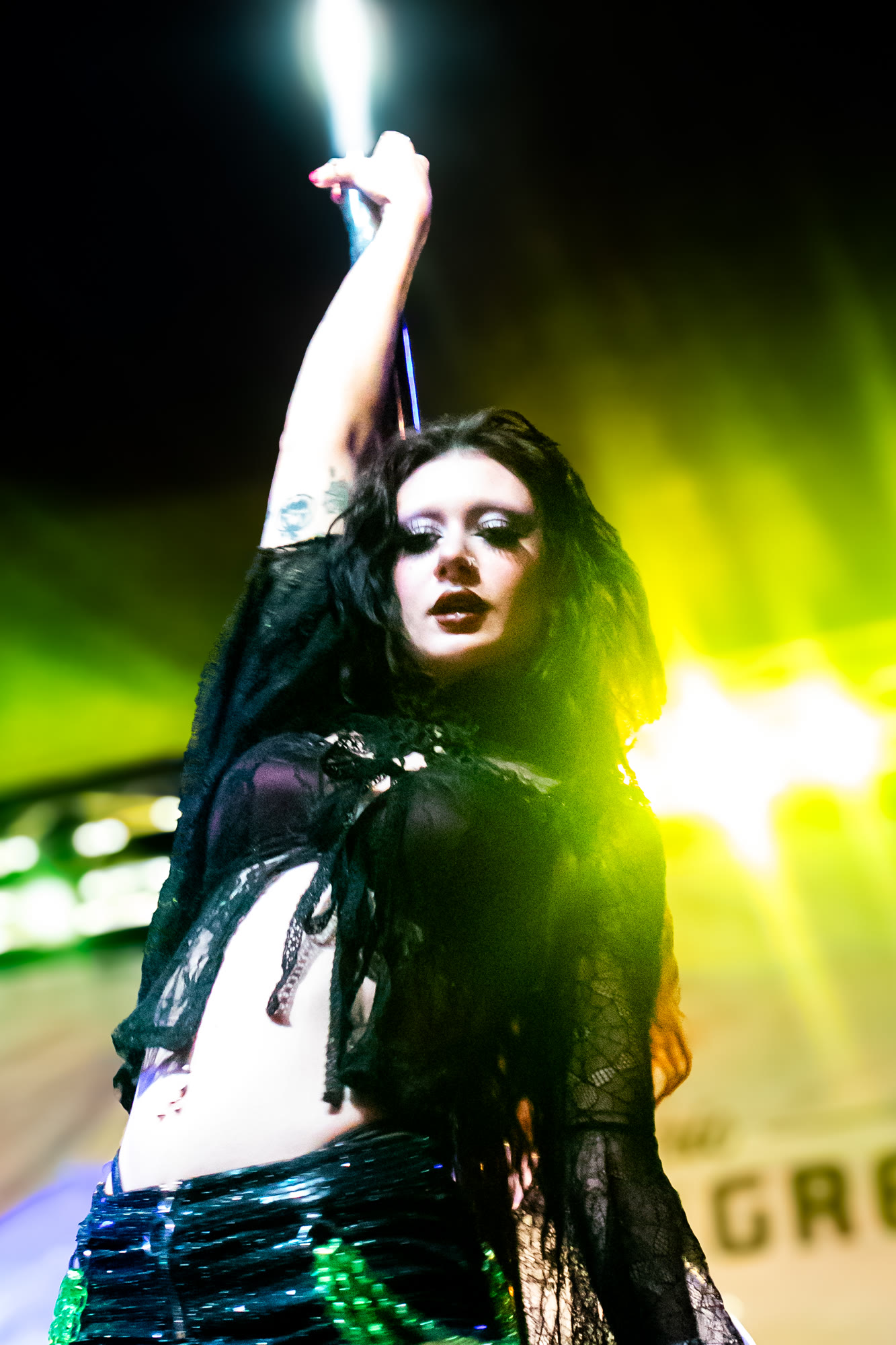 Performer in black lace holds a pole overhead under yellow-green light